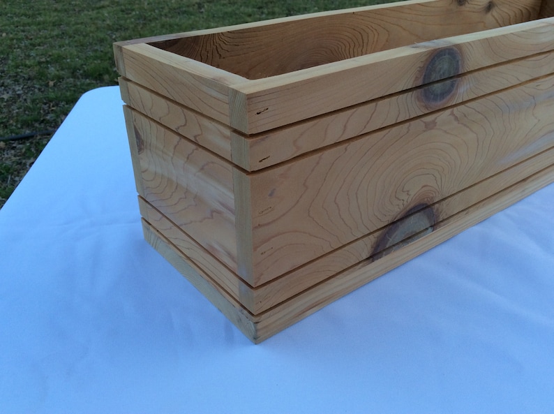 May include: A rectangular wooden planter box with a natural wood finish. The planter has a simple design with three horizontal slats on the front and sides.