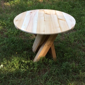 May include: A round wooden table with a natural wood finish. The table has a simple design with a circular top and a tripod base. The table is made of unfinished wood and is sitting on a grassy surface.