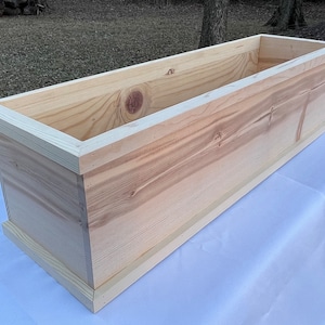 May include: A natural wood planter box with a rectangular shape and a simple design. The planter is made of unfinished wood and has a smooth surface.