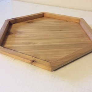 May include: A wooden tray with a unique pentagonal shape. The tray is made of light-colored wood with a natural grain pattern. The edges are slightly raised, creating a defined border. This tray is suitable for serving or decorative purposes.