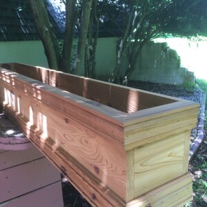 Puede incluir: Una jardinera de madera con un borde decorativo. La jardinera est&aacute; hecha de madera natural con un acabado marr&oacute;n claro. La jardinera est&aacute; sentada sobre una superficie de madera.