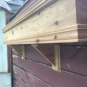 Puede incluir: Un estante de madera con un borde decorativo, montado en una pared de madera roja. El estante est&aacute; soportado por dos soportes triangulares de madera.