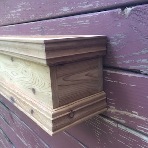Puede incluir: Una jardinera de madera con un acabado natural, montada en una pared rosa desgastada. La jardinera tiene un borde decorativo.