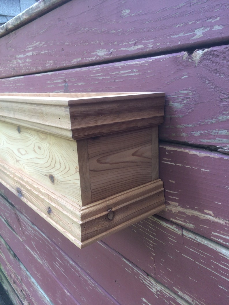 Puede incluir: Una jardinera de madera con un acabado natural, montada en una pared rosa desgastada. La jardinera tiene un borde decorativo.