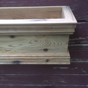 Puede incluir: Una jardinera de madera para ventana con un borde decorativo. La jardinera est&aacute; hecha de madera natural con un acabado marr&oacute;n claro. La jardinera est&aacute; montada en una pared de madera roja.