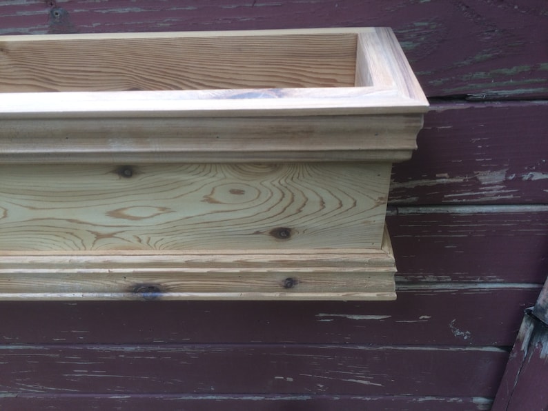 Puede incluir: Una jardinera de madera para ventana con un borde decorativo. La jardinera est&aacute; hecha de madera natural con un acabado marr&oacute;n claro. La jardinera est&aacute; montada en una pared de madera roja.