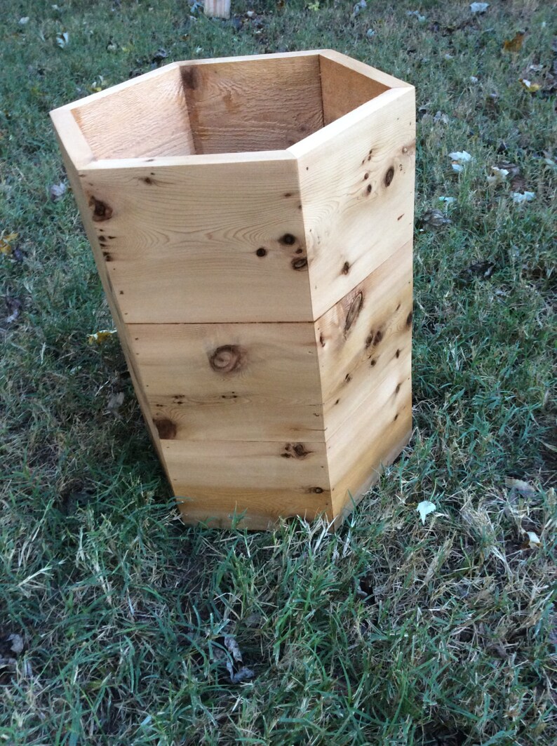 Puede incluir: Un macetero hexagonal de madera natural con un acabado r&uacute;stico. El macetero est&aacute; vac&iacute;o y se encuentra sobre un lecho de hierba verde.