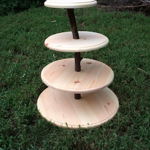 May include: A four-tier wooden cake stand with a natural wood finish. The stand is made of round wooden tiers stacked on top of each other, connected by a natural branch.