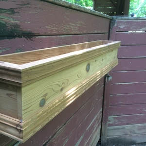 Puede incluir: Una jardinera de madera con un acabado natural est&aacute; montada en una pared de madera roja desgastada. La jardinera tiene un borde decorativo.