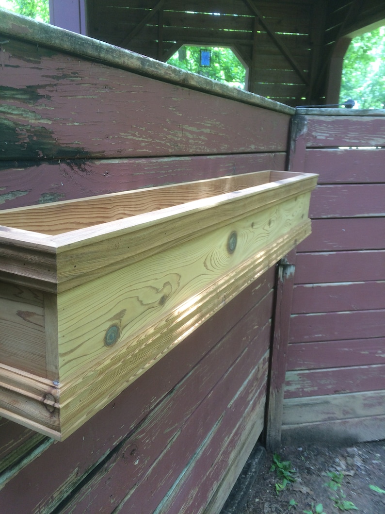 Puede incluir: Una jardinera de madera con un acabado natural est&aacute; montada en una pared de madera roja desgastada. La jardinera tiene un borde decorativo.