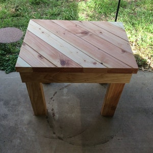 May include: A small wooden table with a chevron pattern top made from light and dark wood planks. The table has four legs and is sitting on a concrete surface.