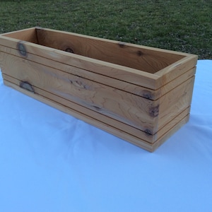 May include: A long, rectangular wooden planter box with a natural wood finish. The planter has a simple design with straight edges and a smooth surface.