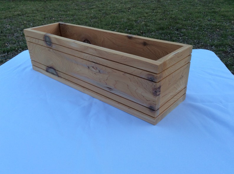 May include: A long, rectangular wooden planter box with a natural wood finish. The planter has a simple design with straight edges and a smooth surface.
