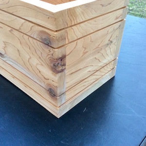 May include: Close-up of the corner of a wooden planter box. The box is made of light-colored wood with a natural finish. The wood grain is visible and the box has a simple, rectangular shape.