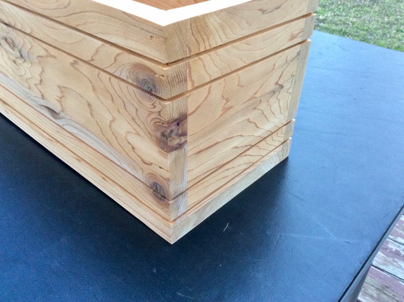 May include: Close-up of the corner of a wooden planter box. The box is made of light-colored wood with a natural finish. The wood grain is visible and the box has a simple, rectangular shape.