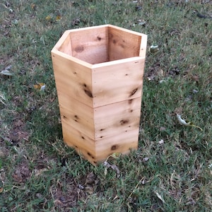 Puede incluir: Un macetero hexagonal de madera natural de color marr&oacute;n claro. El macetero est&aacute; sentado en un lecho de hierba verde.
