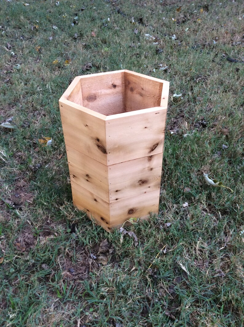 Puede incluir: Un macetero hexagonal de madera natural de color marr&oacute;n claro. El macetero est&aacute; sentado en un lecho de hierba verde.