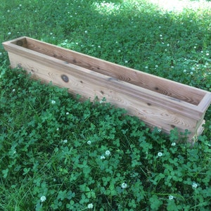 Puede incluir: Una jardinera de madera rectangular y larga, hecha de madera natural con un acabado marr&oacute;n claro. La jardinera est&aacute; sentada sobre un lecho de hierba verde con flores de tr&eacute;bol blanco.