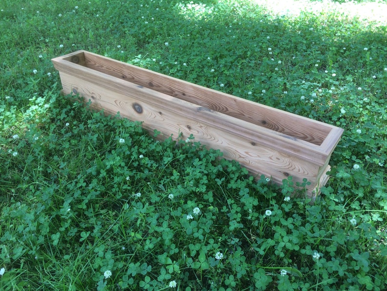 Puede incluir: Una jardinera de madera rectangular y larga, hecha de madera natural con un acabado marr&oacute;n claro. La jardinera est&aacute; sentada sobre un lecho de hierba verde con flores de tr&eacute;bol blanco.