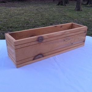May include: A rectangular wooden planter box made of natural cedar wood. The planter has a light brown color and is made of unfinished wood.
