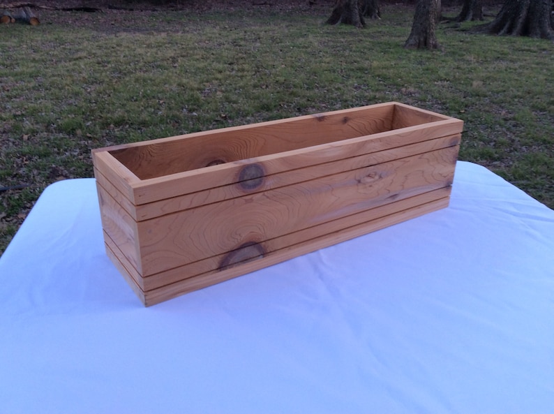 May include: A rectangular wooden planter box made of natural cedar wood. The planter has a light brown color and is made of unfinished wood.