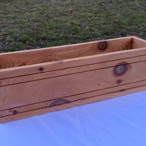 May include: A long, rectangular wooden planter box with a natural wood finish. The planter has a simple design with straight edges and a smooth surface. The wood grain is visible and adds a rustic touch.