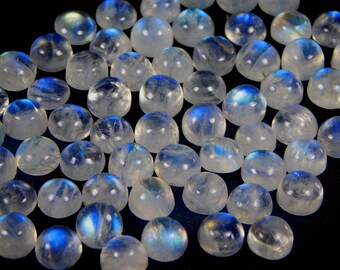 Puede incluir: Un conjunto de piedras preciosas de cabujón de piedra lunar pulidas. Las piedras son translúcidas con una base blanca lechosa y exhiben adularescencia, con destellos azules e iridiscentes. Las piedras preciosas están dispuestas sobre una superficie negra.