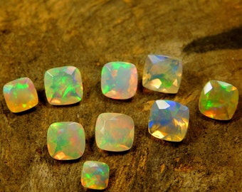 May include: A collection of nine cushion-cut opals, ranging in color from white to green and orange, displayed on a wooden surface.