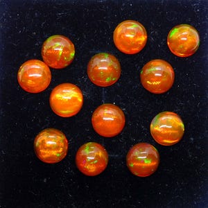 May include: A collection of eleven round, polished opal gemstones. The opals are a vibrant orange color with flashes of green and yellow, set against a black background. These gemstones are ideal for jewelry making.