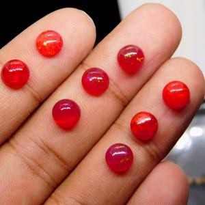 May include: A collection of eight small, round, red gemstones. The gems are displayed on a light-colored surface, with the light reflecting off their polished surfaces. The gemstones have a vibrant, translucent appearance.