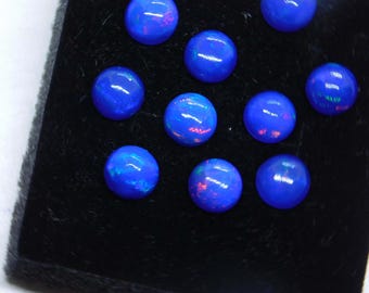May include: A collection of ten round, polished blue opals. Each opal displays a vibrant, iridescent play of color, with flashes of red and green visible within the deep blue hue. The opals are arranged on a black surface.
