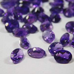 May include: A collection of loose amethyst gemstones, faceted and oval shaped, in various shades of purple. The gemstones are scattered on a white surface.