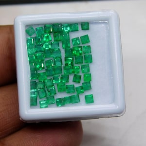May include: A selection of small, square-cut emerald gemstones in a white plastic container. The gemstones are a vibrant green colour and are arranged in a scattered pattern. The container is held by a person's hand.