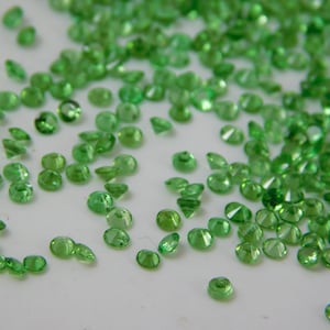 May include: A scattering of small, round, green gemstones on a white surface. The gemstones are faceted and have a shiny, reflective surface.