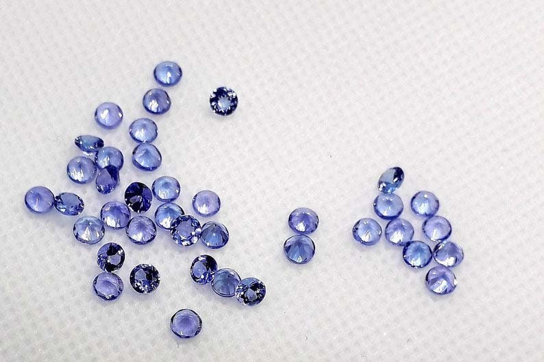 May include: A collection of small, round, light blue gemstones. The gemstones are scattered on a white surface.