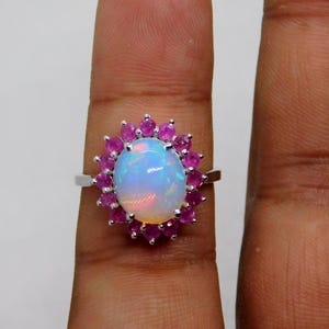 Può includere: Un anello d'argento con un'opale ovale, circondata da piccole pietre rosa rotonde. L'opale ha una base bianca lattea con riflessi iridescenti. Le pietre rosa sono ravvicinate, creando un contrasto vibrante. Gioiello.