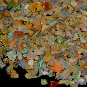 Puede incluir: Vista de cerca de un montón de piedras preciosas de ópalo en bruto. Las piedras muestran una variedad de colores, incluyendo naranja, azul, verde y blanco, con destellos de iridiscencia. La textura rugosa de las piedras es visible.
