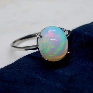 May include: A silver ring featuring a large, oval opal gemstone. The opal displays a vibrant play of color, with flashes of blue, green, and pink. The ring's band is a simple, polished silver.