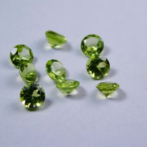 May include: A group of ten round, faceted, green peridot gemstones on a white background.