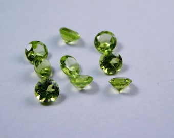 May include: A collection of faceted, round-cut peridot gemstones. The gems are a vibrant, translucent green and are scattered across a white surface. The peridots vary in size and are displayed from multiple angles.