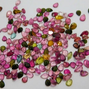 Puede incluir: Una colección de piedras preciosas en forma de pera de varios colores, incluyendo rosa, verde, amarillo y azul. Las piedras preciosas están esparcidas sobre una superficie blanca.