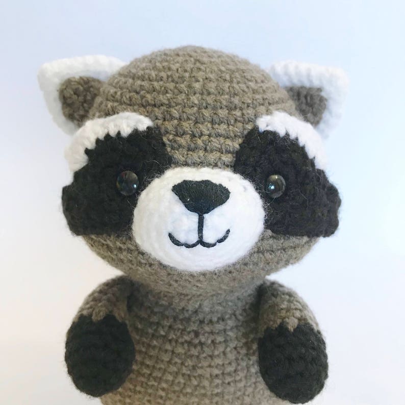 May include: A crocheted raccoon plush toy with black eyes, a white nose and mouth, and black paws. The raccoon is gray with white accents around the eyes and ears.