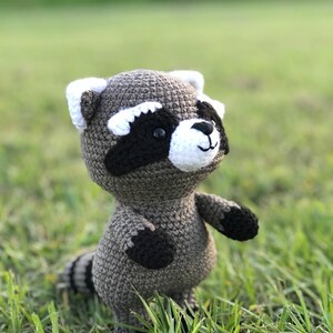 May include: A crocheted raccoon plush toy with black eyes, a white nose, and black and white markings on its face. The toy is standing on green grass.