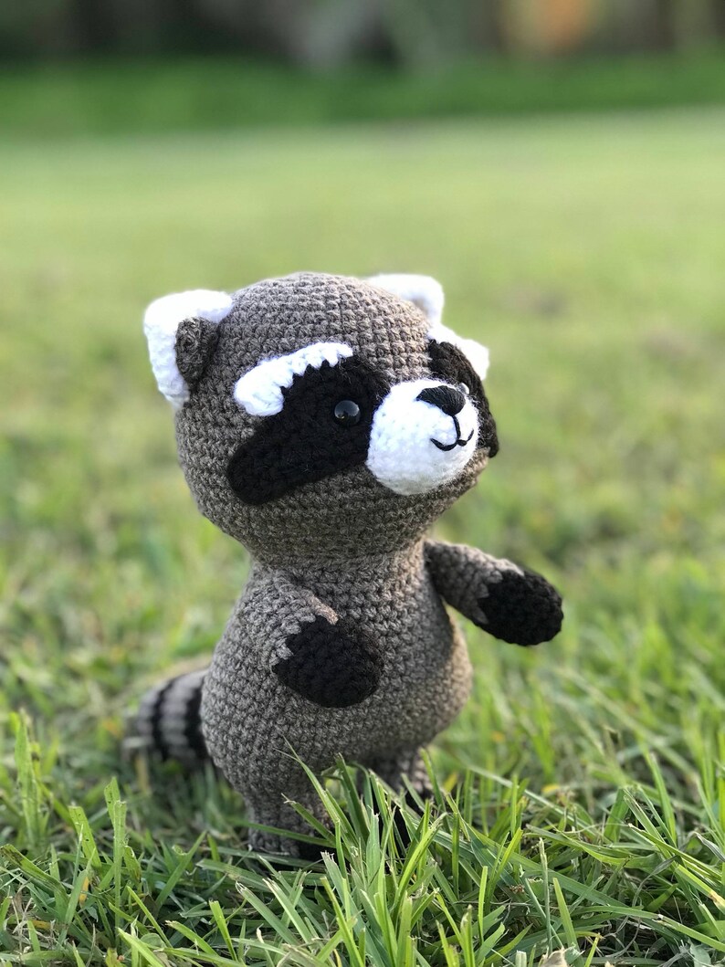 May include: A crocheted raccoon plush toy with black eyes, a white nose, and black and white markings on its face. The toy is standing on green grass.