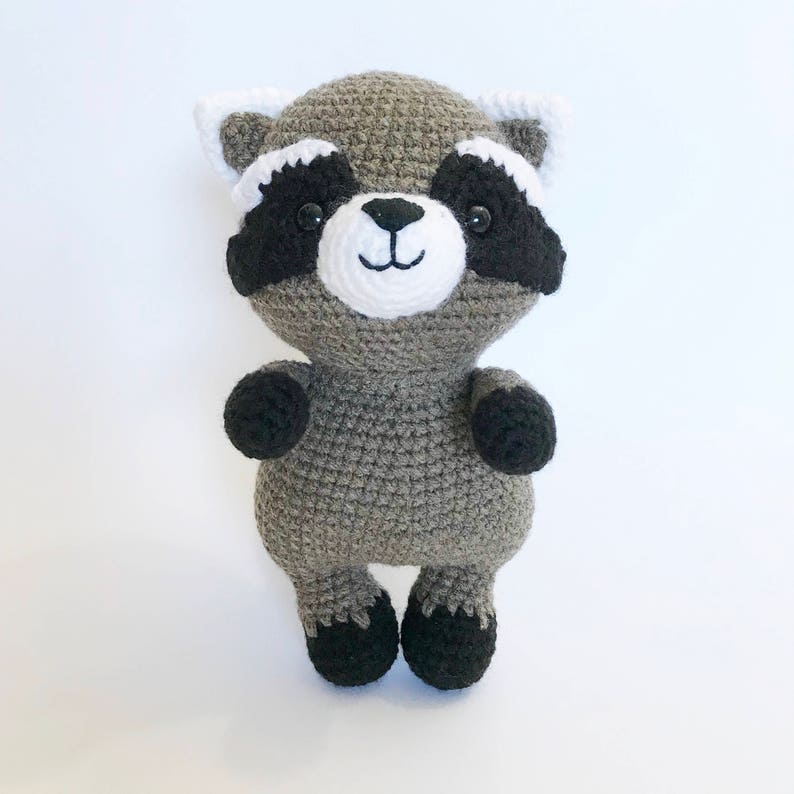 May include: A crocheted raccoon plush toy with black eyes, a white nose, and black and white markings around the eyes. The toy is gray and has black feet.