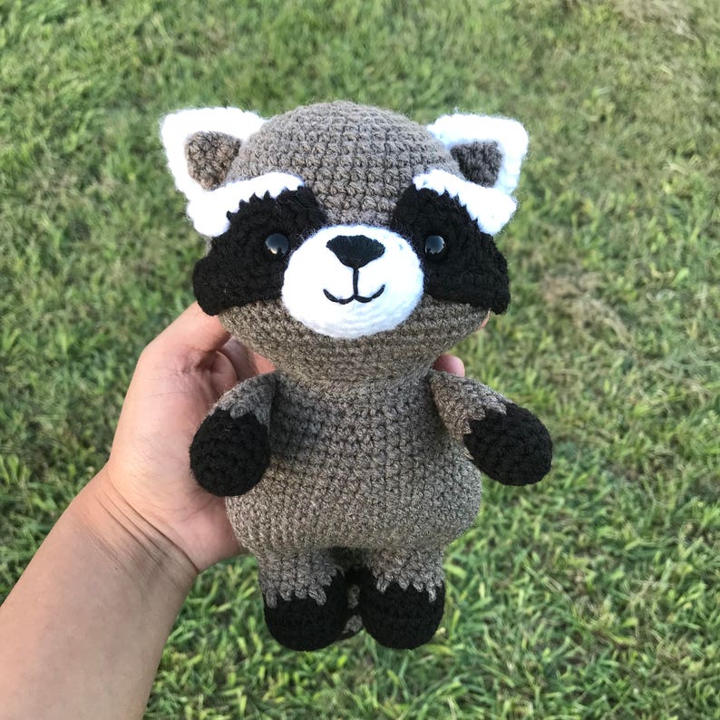May include: A crocheted raccoon plush toy with black eyes, a white nose, and black and white markings around the eyes. The toy is brown and gray and has black feet.