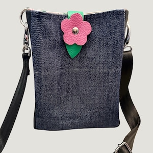 May include: A denim crossbody bag with a pink flower and green leaf accent. The bag has a black adjustable strap.