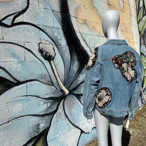 May include: A light blue denim jacket with floral patches on the back and sleeves. The jacket is on a mannequin in front of a brick wall with a mural of large white flowers.