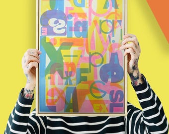 May include: A framed abstract art print with a colourful, layered design. The artwork features overlapping letters and shapes in shades of pink, blue, orange, and green against a yellow background. The frame is a light wood colour.
