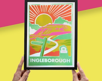 May include: A green and pink illustrated print of Ingleborough, a mountain in the Yorkshire Dales, with a sunburst in the background. The print is framed in black.